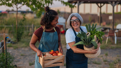 Two women in aprons smiling, holding freshly harvested vegetables outdoors.